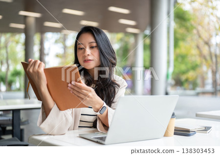 Asian woman reading a book aside laptop and coffee cup while sitting at table in university building Asian woman reading a book aside laptop and coffee cup while sitting at table in university building 138303453