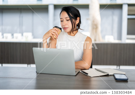Mature asian woman drinking coffee looking at laptop aside book as sitting at wooden table in cafe. Mature asian woman drinking coffee looking at laptop aside book as sitting at wooden table in cafe. 138303484
