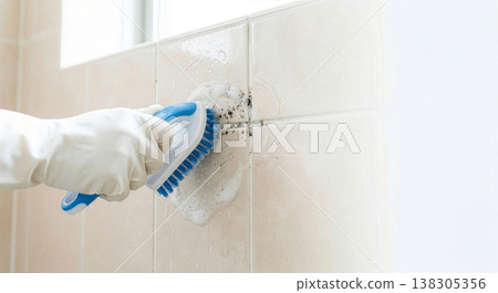 Close-up of a hand wearing rubber gloves cleaning black mold from bathroom tiles with a brush and foam. 138305356