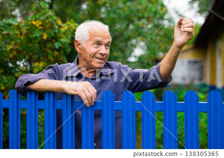 Old elderly farmer stands at fence of his country house and threatens someone with his fist 138305365