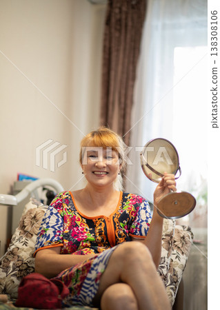 Middle aged woman sitting in chair holding hand mirror. Mature asian woman smiling while checking appearance. Beauty, skincare and self care routine at home. Daily lifestyle concept. 138308106