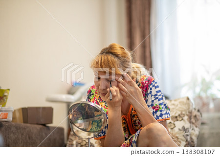 Middle aged woman applying eyeliner looking into mirror. Mature lady wearing colorful blouse doing makeup in living room. Daily skin care and beauty routine for aging skin. Natural look. Middle aged woman applying eyeliner looking into mirror. Mature lady wearing colorful blouse doing makeup in living room. Daily skin care and beauty routine for aging skin. Natural look. 138308107