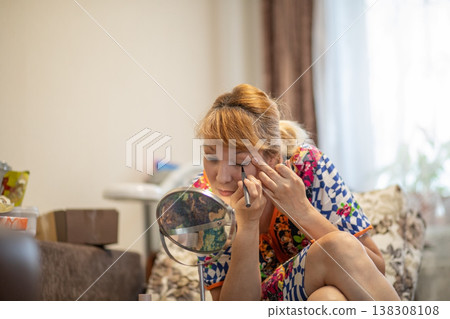 Middle aged woman applying eyeliner looking at small mirror in living room. Daily morning beauty makeup routine of 57 year old lady. Self care and aging gracefully concept at home. 138308108