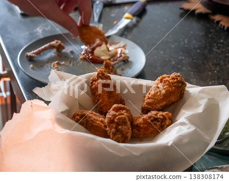 Man eating deep-fried chicken wings from a paper basket. Crispy appetizer served at home. Casual meal, hungry person enjoying fast food snack. Delicious culinary experience and diet concept. Man eating deep-fried chicken wings from a paper basket. Crispy appetizer served at home. Casual meal, hungry person enjoying fast food snack. Delicious culinary experience and diet concept. 138308174