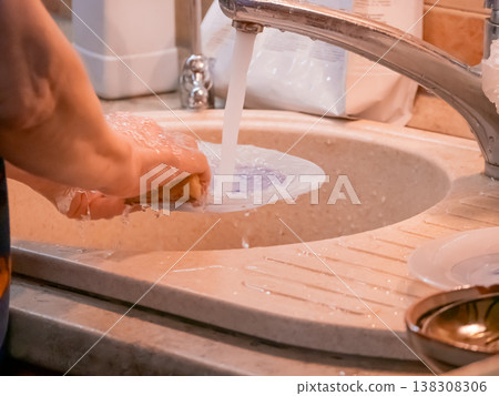 Woman washing plate in kitchen sink. Doing household chores with sponge and running water. Daily domestic routine, cleaning dinnerware after meal. Concept of cleanliness and house care. Woman washing plate in kitchen sink. Doing household chores with sponge and running water. Daily domestic routine, cleaning dinnerware after meal. Concept of cleanliness and house care. 138308306