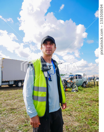 Safety officer man wearing yellow reflective vest and cap standing outdoors at event site. Professional marshal managing logistics and security during outdoor sport competition. Safety officer man wearing yellow reflective vest and cap standing outdoors at event site. Professional marshal managing logistics and security during outdoor sport competition. 138308694