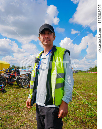Man marshal standing on field during motorcycle race. Security officer wearing safety vest and cap at sport event. Event organizer managing outdoor competition outdoor logistics. Man marshal standing on field during motorcycle race. Security officer wearing safety vest and cap at sport event. Event organizer managing outdoor competition outdoor logistics. 138308695