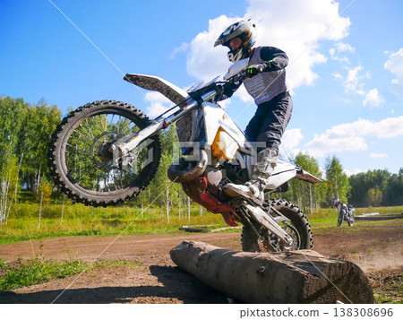 Man motorcyclist performing wheelie on dirt bike over log on off-road track. Professional motocross rider jumping during extreme sports race. Action athlete training in nature wilderness. Man motorcyclist performing wheelie on dirt bike over log on off-road track. Professional motocross rider jumping during extreme sports race. Action athlete training in nature wilderness. 138308696