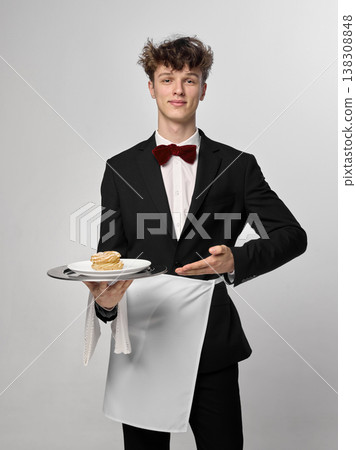 Smiling waiter holding dessert and gesturing toward plate in clean studio portrait. Smiling waiter holding dessert and gesturing toward plate in clean studio portrait. 138308848