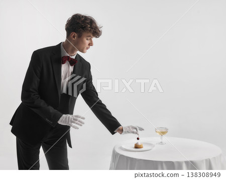 Waiter in formal uniform placing small dessert with cherry on plate on table. Waiter in formal uniform placing small dessert with cherry on plate on table. 138308949