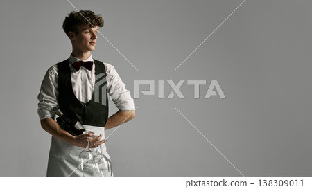 Young waiter in formal uniform holding wine and glasses looking away with confident posture. Young waiter in formal uniform holding wine and glasses looking away with confident posture. 138309011