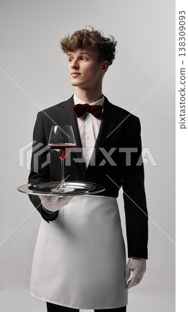 Waiter in formal uniform presenting glass of red wine on tray in studio. Waiter in formal uniform presenting glass of red wine on tray in studio. 138309093