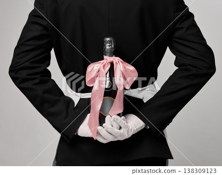 Waiter hiding champagne bottle with ribbon behind back in minimal studio composition. Waiter hiding champagne bottle with ribbon behind back in minimal studio composition. 138309123
