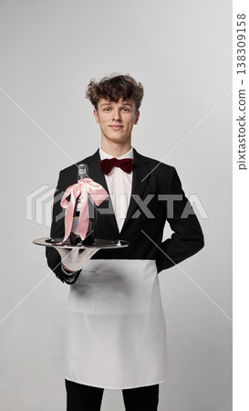 Young waiter presenting champagne bottle decorated with pink ribbon on tray in clean studio Young waiter presenting champagne bottle decorated with pink ribbon on tray in clean studio 138309158
