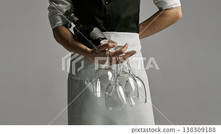 Close up of waiter hands holding wine bottle and empty glasses in minimal studio lighting. Close up of waiter hands holding wine bottle and empty glasses in minimal studio lighting. 138309188