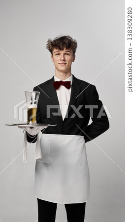Young waiter presenting glass of beer on tray while looking at camera in clean studio Young waiter presenting glass of beer on tray while looking at camera in clean studio 138309280