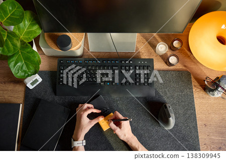 Top view of hands writing on sticky note at desk with monitor, keyboard, mouse and plant Top view of hands writing on sticky note at desk with monitor, keyboard, mouse and plant 138309945