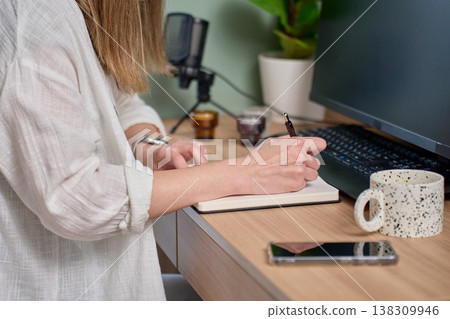 Woman writing in notebook at desk with monitor, keyboard, smartphone and microphone Woman writing in notebook at desk with monitor, keyboard, smartphone and microphone 138309946