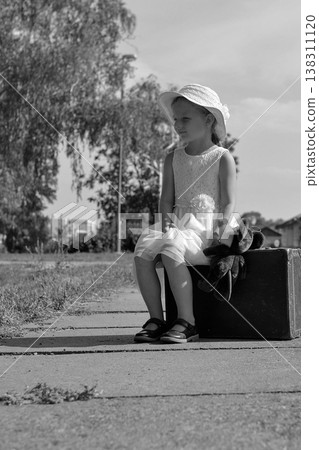 Young girl in a hat and dress sits on a vintage suitcase with a toy, waiting patiently Young girl in a hat and dress sits on a vintage suitcase with a toy, waiting patiently 138311120