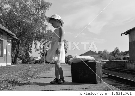 Young girl in a dress and hat waits with a suitcase on a train platform 138311761
