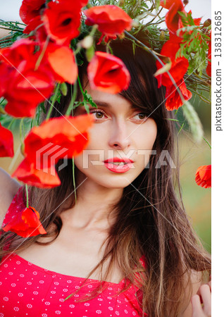 A vertical extreme close-up portrait of a beautiful young woman with long dark hair and red lipstick, whose face is artistically framed by the vibrant red petals of a poppy wreath 138312685