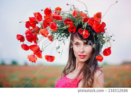 A horizontal portrait of a beautiful young woman with long dark hair, wearing a red patterned dress and an elaborate, asymmetrical wreath of red poppies that cascades to one side, posing in a field at 138312686