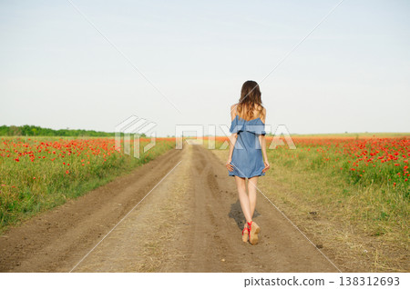 A woman in a blue dress and red sandals walks away on a country road between blooming poppy fields. A woman in a blue dress and red sandals walks away on a country road between blooming poppy fields. 138312693