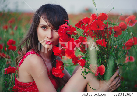 A horizontal close-up portrait of a beautiful young woman with long dark hair and red lipstick, partially hiding her face behind a large bouquet of vibrant red poppies in a blooming field. 138312694