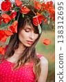A vertical close-up portrait of a beautiful young woman with long dark hair, wearing a red patterned dress and a large wreath of red poppies, 138312695