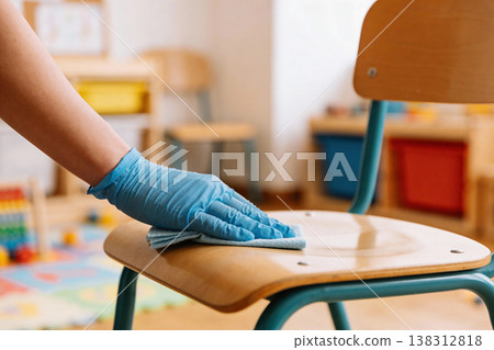 Hand wearing blue glove wipes down a wooden chair seat with a cloth in a classroom 138312818
