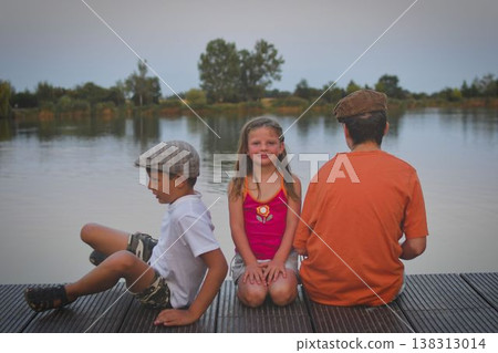 A group of young children sit on a wooden pier by a lake. The young children look toward the future. The concept of summer, holidays, and a carefree childhood. Summer and childhood concept. Children A group of young children sit on a wooden pier by a lake. The young children look toward the future. The concept of summer, holidays, and a carefree childhood. Summer and childhood concept. Children 138313014