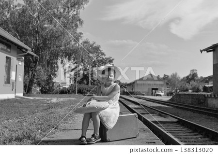 Young girl in a hat and dress sits on a suitcase at a train station platform with tracks 138313020