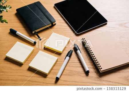 Flat lay of office supplies including notebooks, sticky notes, pens, and a tablet on a wooden desk 138313129