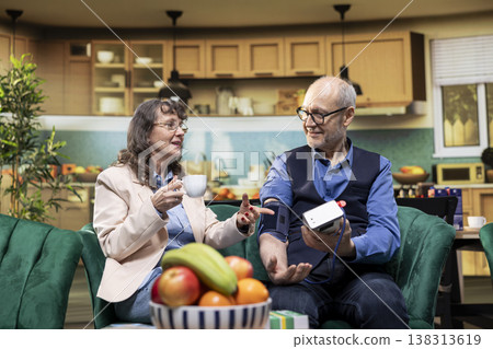 Retired senior people using electronic blood pressure device with cuff and pump, monitoring pulse pressure and vital signs in living room. Daily care supports recovery and wellbeing, quality of life. 138313619