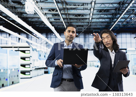 Team of business professionals in microchip factory cleanroom overseeing hardware production metrics. Management executives using devices to monitor manufacturing infrastructure bottlenecks 138313643