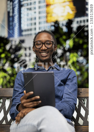 Portrait of cheerful woman using tablet to watch movies, sitting on city park bench. Cheerful relaxed african american person using device, enjoying TV shows entertainment outside 138313689