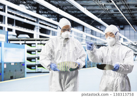 Team of engineers doing research and development on silicon wafer, brainstorming in cleanroom. Teamworking colleagues in protective suit examining microelectronic semiconductor substrate for defects 138313758