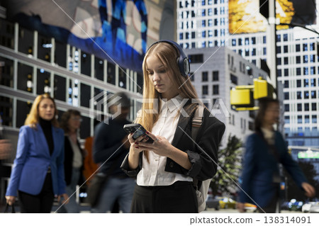 Young adult college student with big headphones browsing smartphone at intersection, checking her messages while waiting at a red light. Hipster girl from gen z walking the street. Young adult college student with big headphones browsing smartphone at intersection, checking her messages while waiting at a red light. Hipster girl from gen z walking the street. 138314091