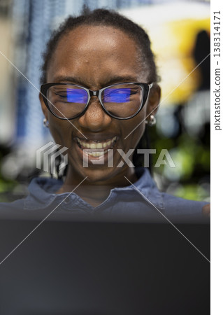 Close up of ecstatic woman laughing, using laptop to watch funny online clips on city park bench. Overjoyed African american girl amused by hilarious memes on notebook, enjoying leisure time outside 138314171