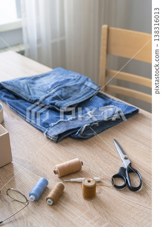 Old denim jeans on a wooden table, surrounded by sewing threads, needles, scissors. Promoting sustainable fashion through craft and repair, upcycling concept Old denim jeans on a wooden table, surrounded by sewing threads, needles, scissors. Promoting sustainable fashion through craft and repair, upcycling concept 138316013