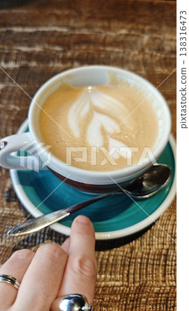 Coffee cup with latte art featuring leaf design on creamy surface, placed on a turquoise saucer with a silver spoon and hand visible in a cozy cafe setting Coffee cup with latte art featuring leaf design on creamy surface, placed on a turquoise saucer with a silver spoon and hand visible in a cozy cafe setting 138316473