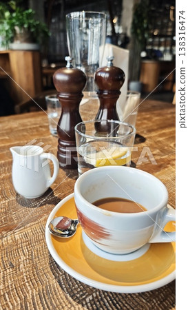 Coffee cup with saucer and spoon on wooden table, accompanied by glass of water with lemon slice and salt and pepper shakers in a cozy restaurant setting Coffee cup with saucer and spoon on wooden table, accompanied by glass of water with lemon slice and salt and pepper shakers in a cozy restaurant setting 138316474
