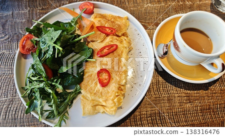 Plate of fluffy omelette garnished with sliced red chili peppers, served alongside fresh salad of arugula and cherry tomatoes, with a cup of coffee on a wooden table Plate of fluffy omelette garnished with sliced red chili peppers, served alongside fresh salad of arugula and cherry tomatoes, with a cup of coffee on a wooden table 138316476