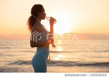 Fit woman drinking water at the beach after running outdoors in the morning. Sporty female relaxing after training. Fit woman drinking water at the beach after running outdoors in the morning. Sporty female relaxing after training. 138316528