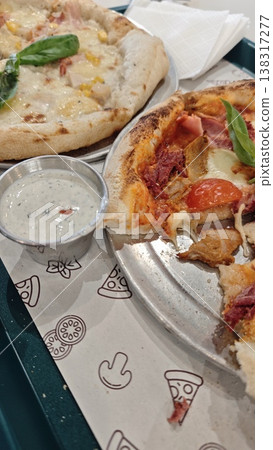 Two pizzas on a tray, one topped with cheese and ham, the other with pepperoni and vegetables, accompanied by a small cup of dipping sauce on a patterned paper 138317277