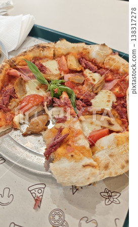 Pizza topped with sliced chicken, tomatoes, and cheese on a metal plate, garnished with fresh basil leaves, served on a patterned table surface Pizza topped with sliced chicken, tomatoes, and cheese on a metal plate, garnished with fresh basil leaves, served on a patterned table surface 138317278