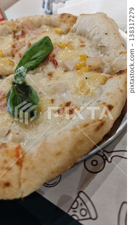 Close-up of freshly baked pizza topped with cheese, corn, chicken, and basil leaves on a round metal plate with a patterned paper background 138317279