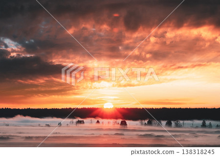 Bright sun setting behind a dark forest over a misty field 138318245