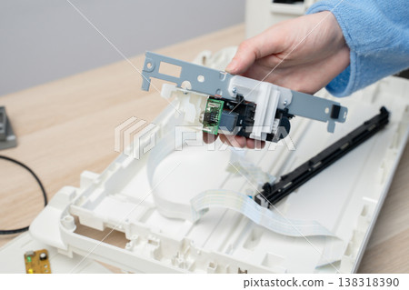 Close-up of a technician's hands repairing a flatbed scanner printer component, showing circuit board, ribbon cable and plastic chassis during electronic maintenance and equipment troubleshooting. Close-up of a technician's hands repairing a flatbed scanner printer component, showing circuit board, ribbon cable and plastic chassis during electronic maintenance and equipment troubleshooting. 138318390