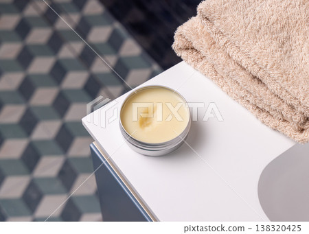 Opened cosmetic lip balm metal tin near beige bath towel in blue tiled bathroom top view 138320425